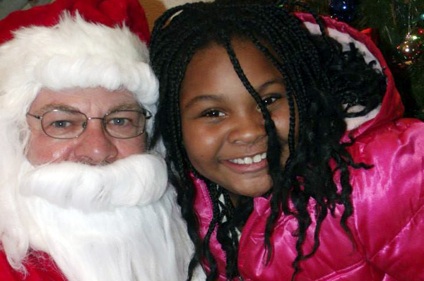 Picture of a pretty pre-teen African American girl in pink jacket sitting with volunteer Santa